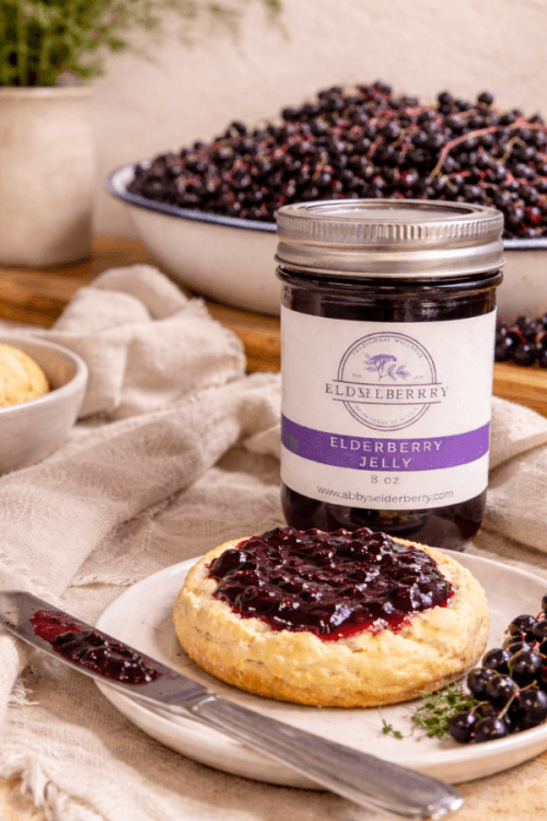 Abby's Elderberry jelly spread on a biscuit beside a jar and a bowl of elderberries